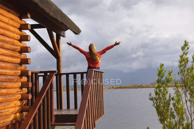 Rückansicht einer kaukasischen Frau, die eine gute Reise in die Berge unternimmt, auf einem Balkon in einer Hütte steht und die Hände in die Luft reckt — Stockfoto