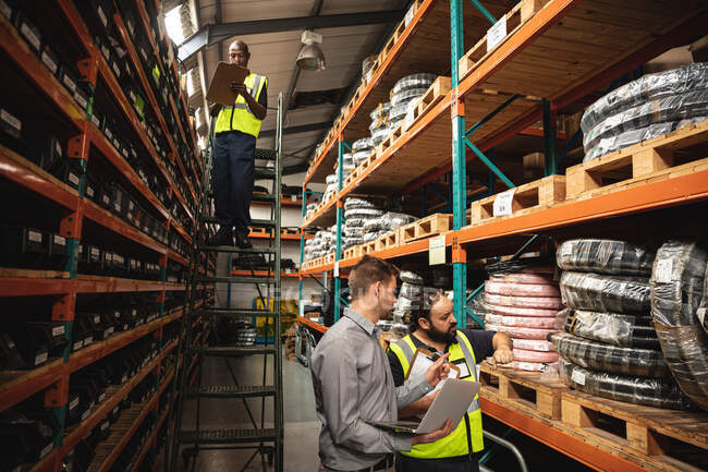 African American and Caucasian factory workers wearing a high vis vest and Caucasian male supervisor using laptop and checking stock. Workers in industry at a factory making hydraulic equipment. — Stock Photo