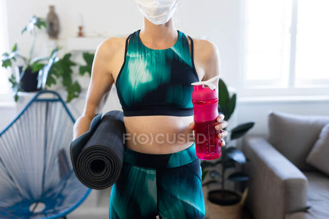 Mid section of woman spending time at home, wearing sportswear and a face mask against coronavirus, covid 19, holding a mat and a plastic bottle. Social distancing and self isolation in quarantine lockdown. — Stock Photo