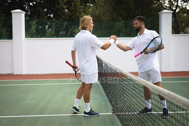 Белый и смешанная раса мужчин в теннисных белых проводить время на корте вместе, играть в теннис в солнечный день, пожимая руки, держа теннисные ракетки — стоковое фото