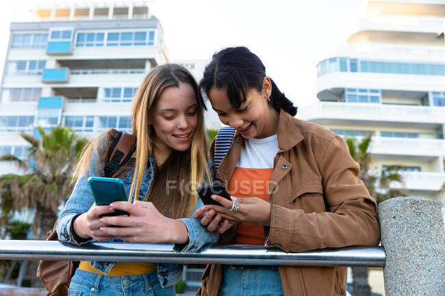 Front view of a Caucasian and a mixed race girls enjoying time hanging out together on a sunny day, standing and leaning on the railing, holding and looking at their smartphones. — Stock Photo
