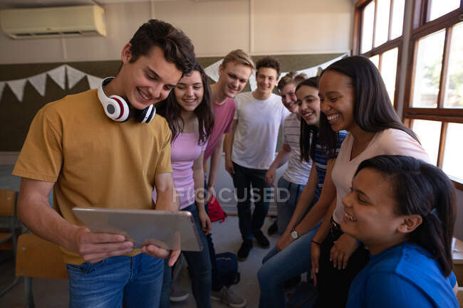Front view of a multi-ethnic group of teenage school pupils standing together in a classroom looking at a tablet computer and smiling at break time — Stock Photo