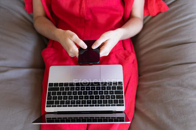 Средняя часть женщины проводит время дома, одетая в розовое платье, сидя на диване и пользуясь ноутбуком и смартфоном. Социальное дистанцирование и самоизоляция в карантинной изоляции. — стоковое фото