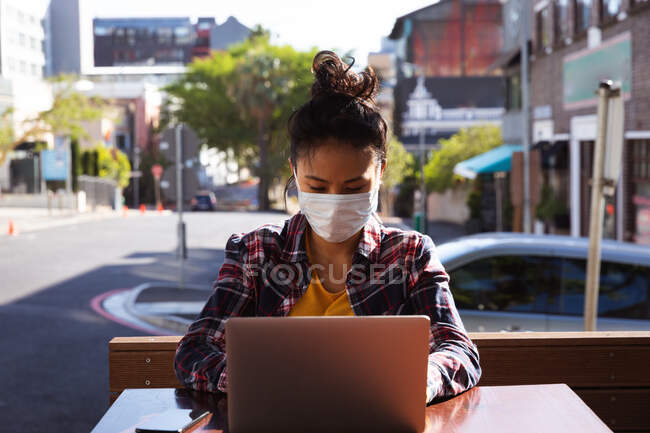 Vorderansicht einer Frau mit langen dunklen Haaren, die tagsüber an einem Tisch in einem Café sitzt, eine Gesichtsmaske gegen Luftverschmutzung und Coronavirus trägt und an einem Laptop mit Gebäuden im Hintergrund arbeitet. — Stockfoto