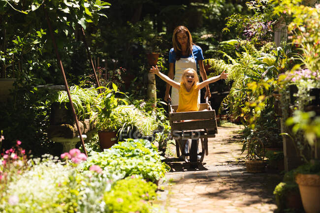 Una mujer caucásica vistiendo un delantal y su hija disfrutando del tiempo en un jardín soleado, la hija sentada en una carretilla mientras la madre la empuja - foto de stock