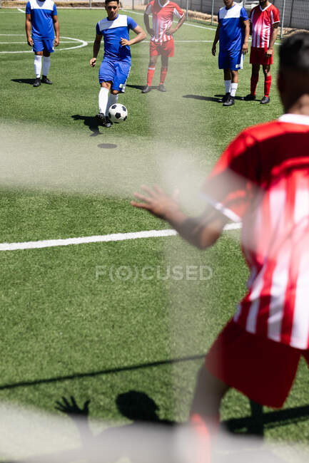 Дві етнічні команди чоловіків-футболістів в командній смузі грають гру на спортивному полі на сонці, один гравець штовхає м'яча воротаря в ворота . — стокове фото