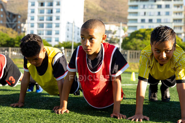 Vista frontal de cerca de un grupo multiétnico de jugadores de fútbol de niños haciendo flexiones de presión en una fila en un campo de juego en el sol durante una sesión de entrenamiento de fútbol - foto de stock