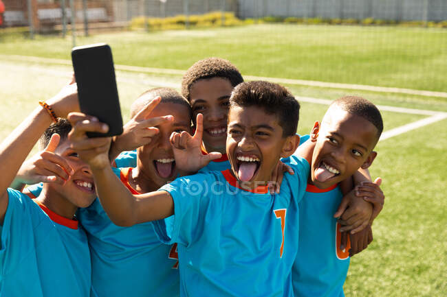 Вид спереди на многоэтническую группу мальчишек-футболистов, одетых в раздевалку своей команды, стоящих на игровом поле, делающих селфи со смартфоном, позирующих с обнимающимися руками, улыбающихся и делающих лица — стоковое фото