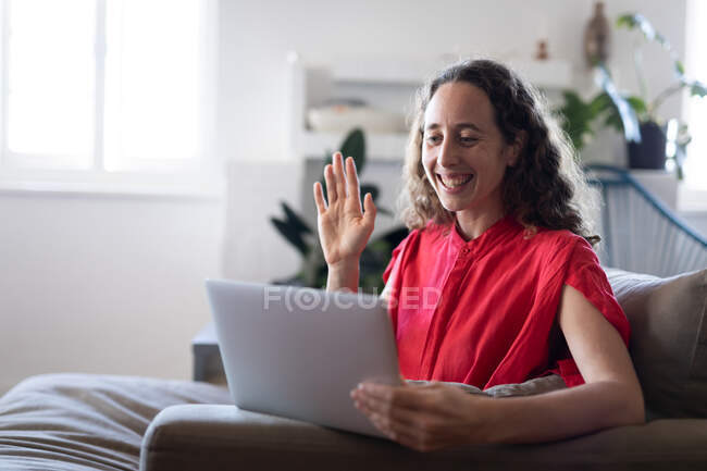 Femme blanche passant du temps à la maison, portant une robe rose, assise sur un canapé et tenant son ordinateur portable, interagissant en utilisant les médias sociaux. Distance sociale et isolement personnel en quarantaine. — Photo de stock