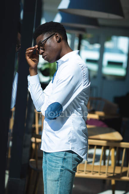 An African American businessman with short hair, wearing a white shirt and glasses standing inside a cafe and looking at the window touching his forehead — Stock Photo