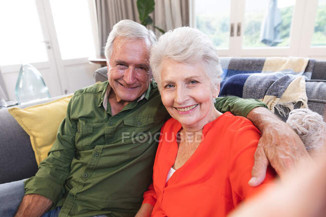 Retrato de um casal caucasiano aposentado feliz em casa em sua sala de estar, sentado em um sofá, ambos olhando para câmera e sorrindo, a mulher tomando uma selfie, casal isolando durante coronavírus covid19 pandemia — Fotografia de Stock