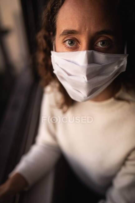 Retrato de una mujer caucásica que pasa tiempo en casa aislándose y distanciándose socialmente en el bloqueo de cuarentena durante la epidemia de coronavirus covid 19, usando una máscara facial contra el coronavirus covid19, mirando directamente a una cámara. - foto de stock