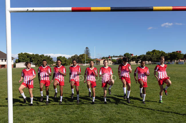 Vista frontal de un equipo masculino multiétnico adolescente de jugadores de rugby que usan su tira de equipo, calentándose en el campo de juego, corriendo en el acto - foto de stock