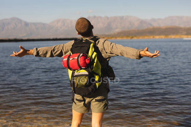 Rückseite Nahaufnahme eines kaukasischen Mannes, der sich bei einem Ausflug in die Berge amüsiert, auf einem Pfad stehend, am Ufer, die Arme weit ausgebreitet, an einem sonnigen Tag — Stockfoto