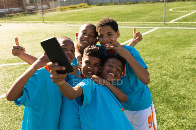 Vista frontal de un grupo de jóvenes futbolistas multiétnicos usando su tira de equipo, de pie en un campo de juego tomando selfie con teléfono inteligente - foto de stock