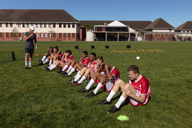 Vue latérale d'une équipe masculine de rugby multi-ethnique adolescente portant leur bande d'équipe, s'échauffant sur le terrain de jeu, faisant des redressements assis, avec leur entraîneur sifflant. — Photo de stock