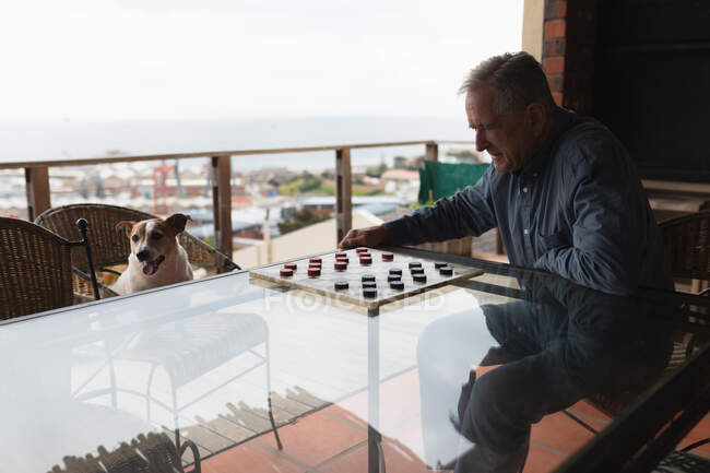 Біля вікна сидить старший кавказький чоловік, який відпочиває вдома, сидячи за столом, граючи з домашнім собакою на стільці навпроти нього. — стокове фото