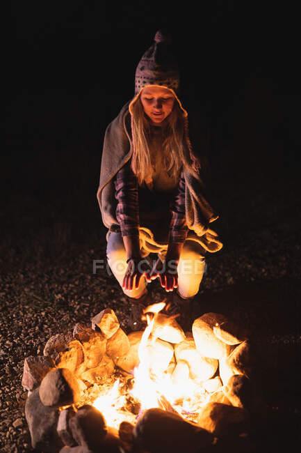Front view of a Caucasian woman having a good time on a trip to the mountains, kneeling by a campfire, warming his hands — Stock Photo