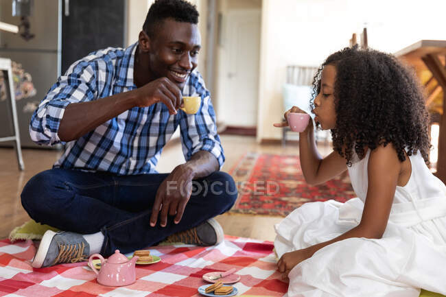 Afro-americano menina social distanciamento em casa durante quarentena bloqueio, brincando com seu pai, tendo uma boneca chá festa . — Fotografia de Stock