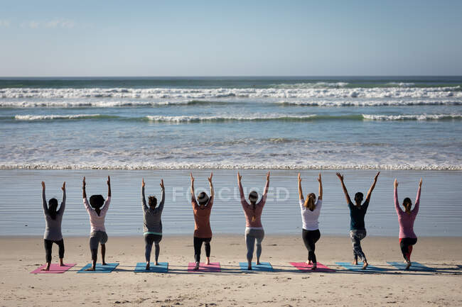 Вид сзади на многонациональную группу подруг, занимающихся спортом на пляже в солнечный день, практикующих йогу, стоящих в позиции йоги. — стоковое фото