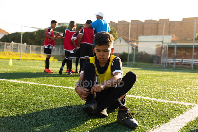 Front view of a mixed race boy soccer player sitting on a playing field in the sun putting on his football boots during a training session, with his teammates listening to their coach in the background — Stock Photo
