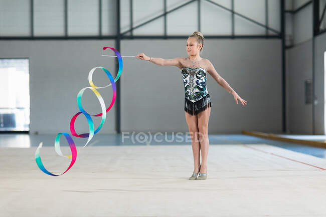 Вид спереди на юную кавказскую гимнастку, выступающую в тренажерном зале, тренирующуюся с ленточкой, носящую разноцветный трико. — стоковое фото