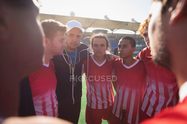 Side view of a multi-ethnic group of male field hokey players and a Caucasian male field hokey coach, preparing before a game, huddling, interacting on a sunny day — Stock Photo
