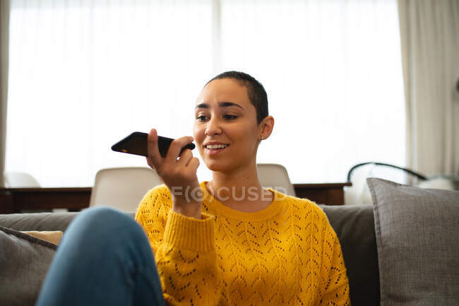 Передній вид змішаної раси жінка розслабляється вдома, сидячи на дивані з ногами вгору, тримаючи смартфон, розмовляючи і посміхаючись — стокове фото