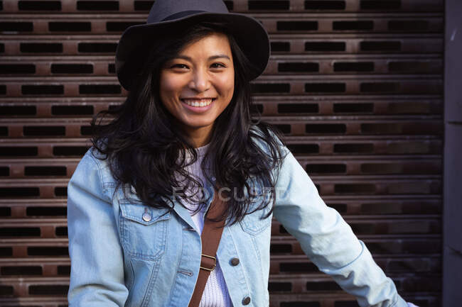 Portrait d'une heureuse femme métissée aux longs cheveux foncés dans les rues de la ville pendant la journée, vêtue d'un chapeau et d'une veste en denim, souriant à la caméra avec un mur en arrière-plan. — Photo de stock