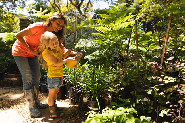 Кавказская женщина и ее дочь наслаждаются временем вместе в солнечном саду, глядя на растения, поливая растения лейкой — стоковое фото