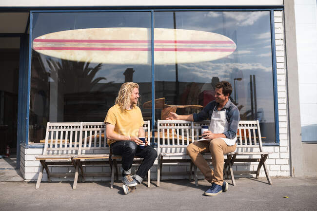 Deux surfeurs masculins caucasiens font une pause pendant leur travail dans leur studio, assis sur un banc et interagissant en buvant un café à emporter et en prenant une collation. — Photo de stock
