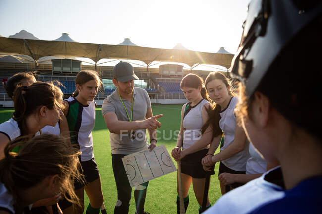 Side view of a group of female Caucasian field hockey players preparing before a game, huddling around their Caucasian male coach, on a hockey pitch, who gives them game plan, and points at something, on a sunny day — Stock Photo