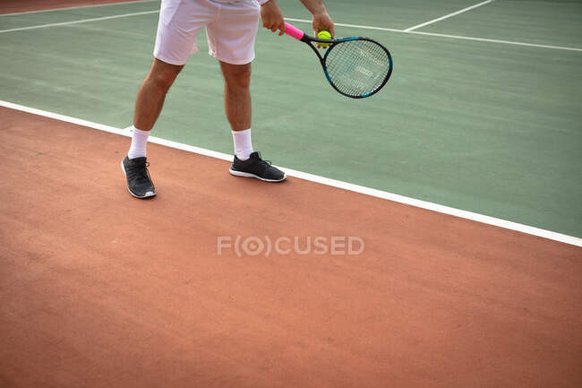 Vista de mitad de la sección del hombre con ropa blanca de tenis pasar tiempo en una cancha jugando al tenis en un día soleado, preparándose para golpear una pelota - foto de stock