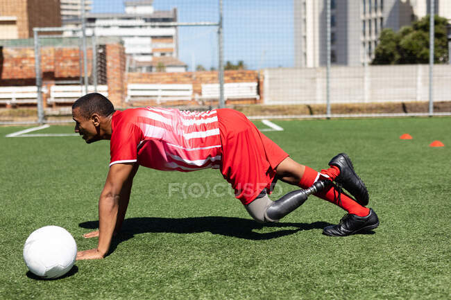 Jogador de futebol masculino de raça mista com perna protética vestindo um treinamento de tira de equipe em um campo de esportes ao sol, aquecendo-se fazendo flexões com bola ao lado dele. — Fotografia de Stock