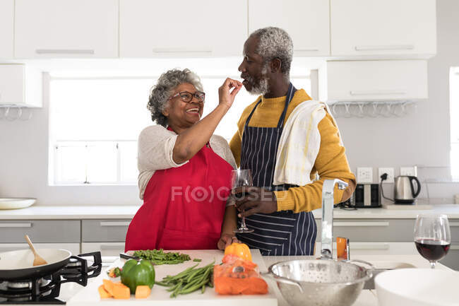 Ein älteres afroamerikanisches Paar, das während der Coronavirus-Epidemie 19 Zeit zu Hause verbringt, soziale Distanzierung und Selbstisolierung in Quarantäne, steht in der Küche und bereitet Essen zu, der Mann hält ein Glas Rotwein — Stockfoto