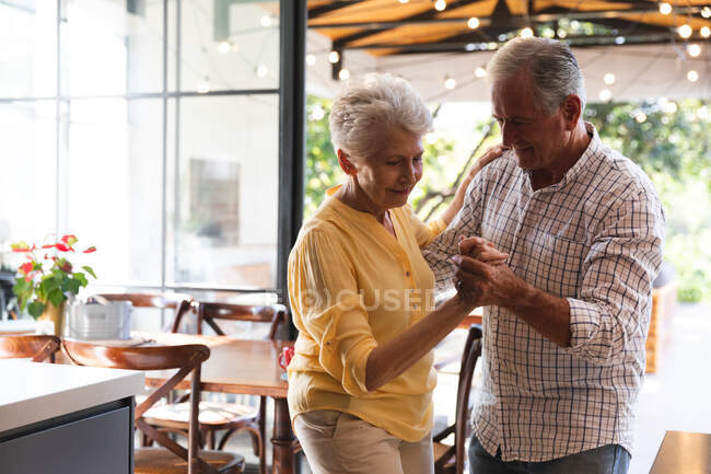 Feliz pareja de ancianos caucásicos jubilados en casa tomados de la mano, bailando juntos en su cocina y sonriendo, en casa juntos aislando durante coronavirus covid19 pandemia - foto de stock