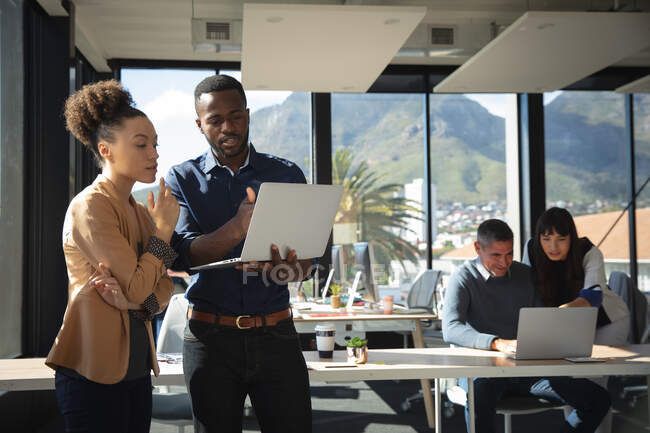 Une femme d'affaires métisse et un homme d'affaires afro-américain travaillant dans un bureau moderne, utilisant un ordinateur portable et parlant, avec leurs collègues travaillant en arrière-plan — Photo de stock