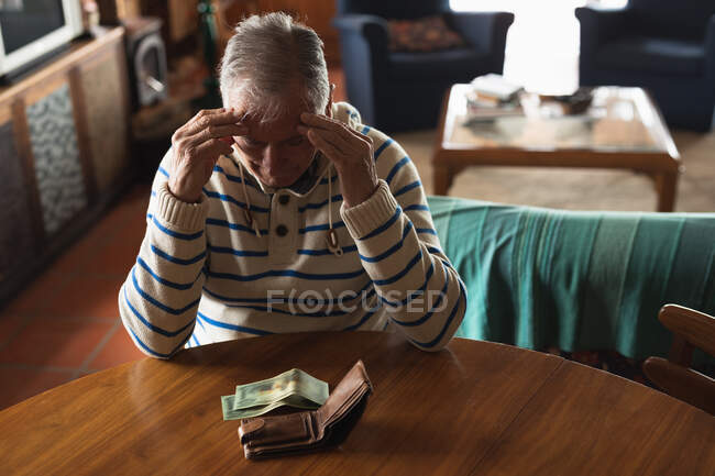 Вид спереди пожилого кавказца дома, сидящего за столом в столовой, смотрящего на деньги в бумажнике, держащего голову и думающего: — стоковое фото