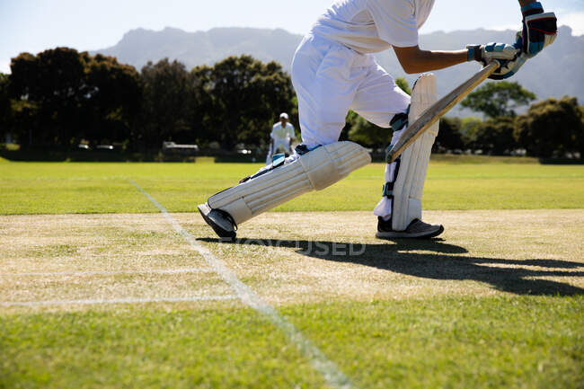 Seitenansicht niedriger Abschnitt eines gemischten männlichen Cricketspielers, der Weiße trägt, einen Cricketschläger in der Hand hält und sich darauf vorbereitet, den Ball zu treffen, auf dem Spielfeld während eines Cricketspiels, während ein anderer Spieler im Hintergrund steht. — Stockfoto