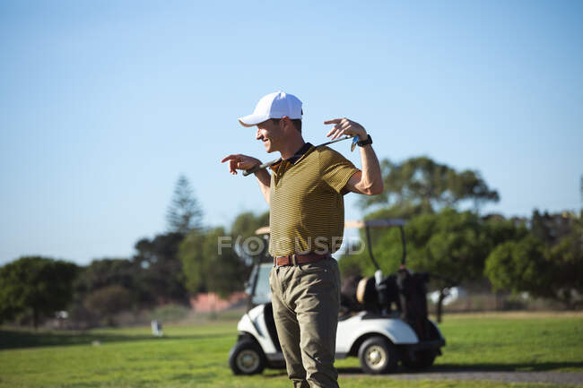 Vue latérale d'un homme caucasien sur un terrain de golf par une journée ensoleillée avec ciel bleu, tenant un club de golf, avec une voiturette de golf en arrière-plan — Photo de stock