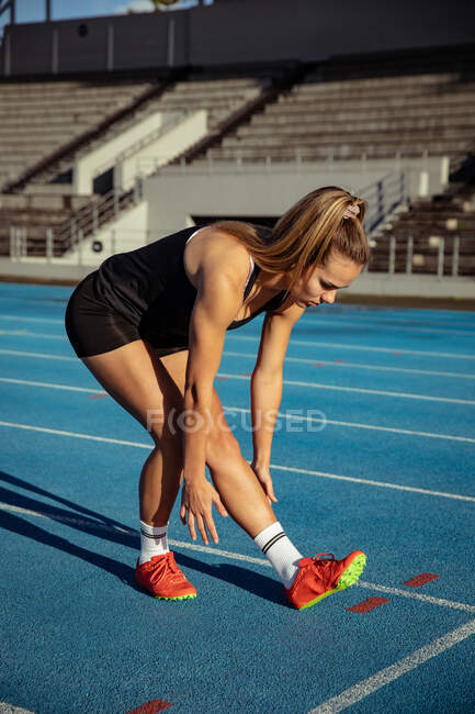 Vista lateral de uma atleta caucasiana praticando em um estádio de esportes, estendendo-se em uma pista de corrida. — Fotografia de Stock