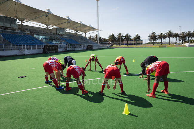 Side view of a multi-ethnic group of teenage male field hockey players and a Caucasian field hockey coach, preparing before a game, working out on a field hokey pitch, doing bend downs in a circle on a sunny day — Stock Photo