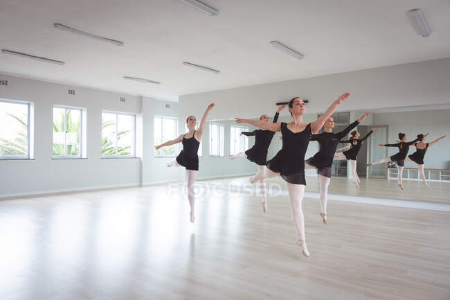 Група кавказьких танцюристок привабливого балету в чорному вбранні під час класу балету в яскравій студії, танцюють і стрибають на одній нозі в унісон.. — стокове фото