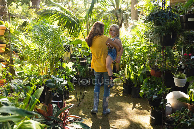 Uma mulher caucasiana e sua filha desfrutando de tempo juntos em um jardim ensolarado, mãe segurando filha em seus braços, a filha olhando para a câmera e sorrindo — Fotografia de Stock