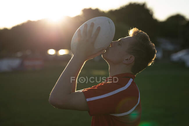 Vista lateral de cerca de un jugador de rugby masculino caucásico adolescente con tira roja del equipo, sosteniendo y besando la pelota de rugby, de pie en un campo de juego después de un partido, retroiluminado por la luz del sol - foto de stock