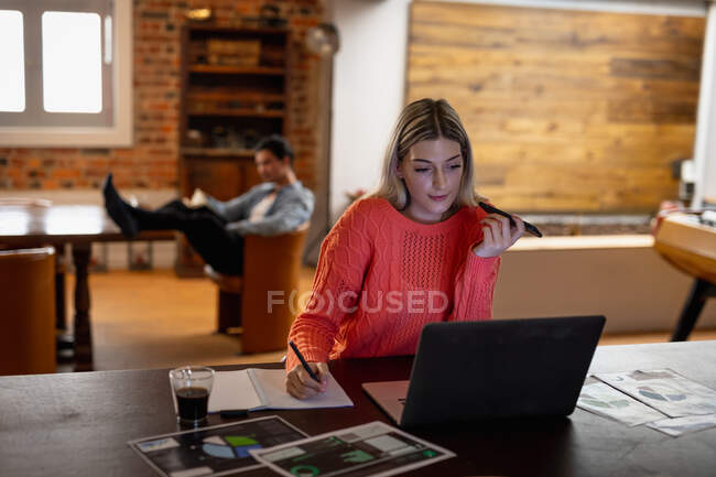 Vue de face d'une jeune femme caucasienne, assise dans le salon, utilisant son ordinateur portable tout en travaillant, son partenaire est assis en arrière-plan. — Photo de stock