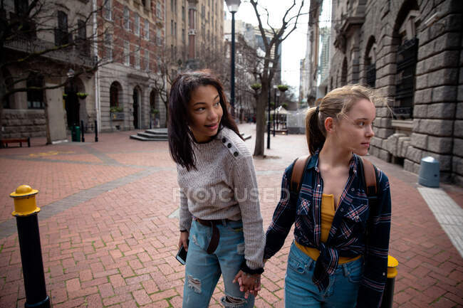 Vista frontale di un caucasico e di una razza mista ragazze godendo il tempo appendere insieme in una giornata di sole, in piedi sul marciapiede, tenendosi per mano. — Foto stock