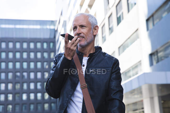Homem caucasiano sênior, vestindo roupas casuais, nas ruas da cidade durante o dia, usando um smartphone . — Fotografia de Stock