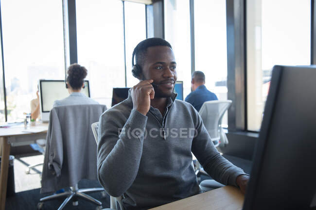 Un hombre de negocios afroamericano que trabaja en una oficina moderna, sentado en un escritorio, usando una computadora, usando auriculares y hablando, con sus colegas trabajando en el fondo - foto de stock