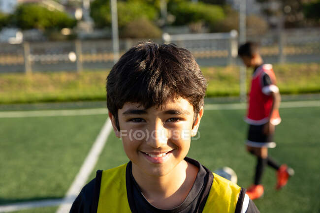 Портрет крупним планом впевнений змішаної раси хлопчик-футболіст в командній смузі, стоячи на ігровому полі на сонці, дивлячись на камеру і посміхаючись, з товаришем по команді на задньому плані — стокове фото
